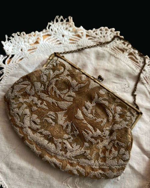 An antique-style evening clutch purse with intricate gold and white floral embroidery, a gold-tone metal frame, and a chain strap, displayed on a white lace doily against a black background.